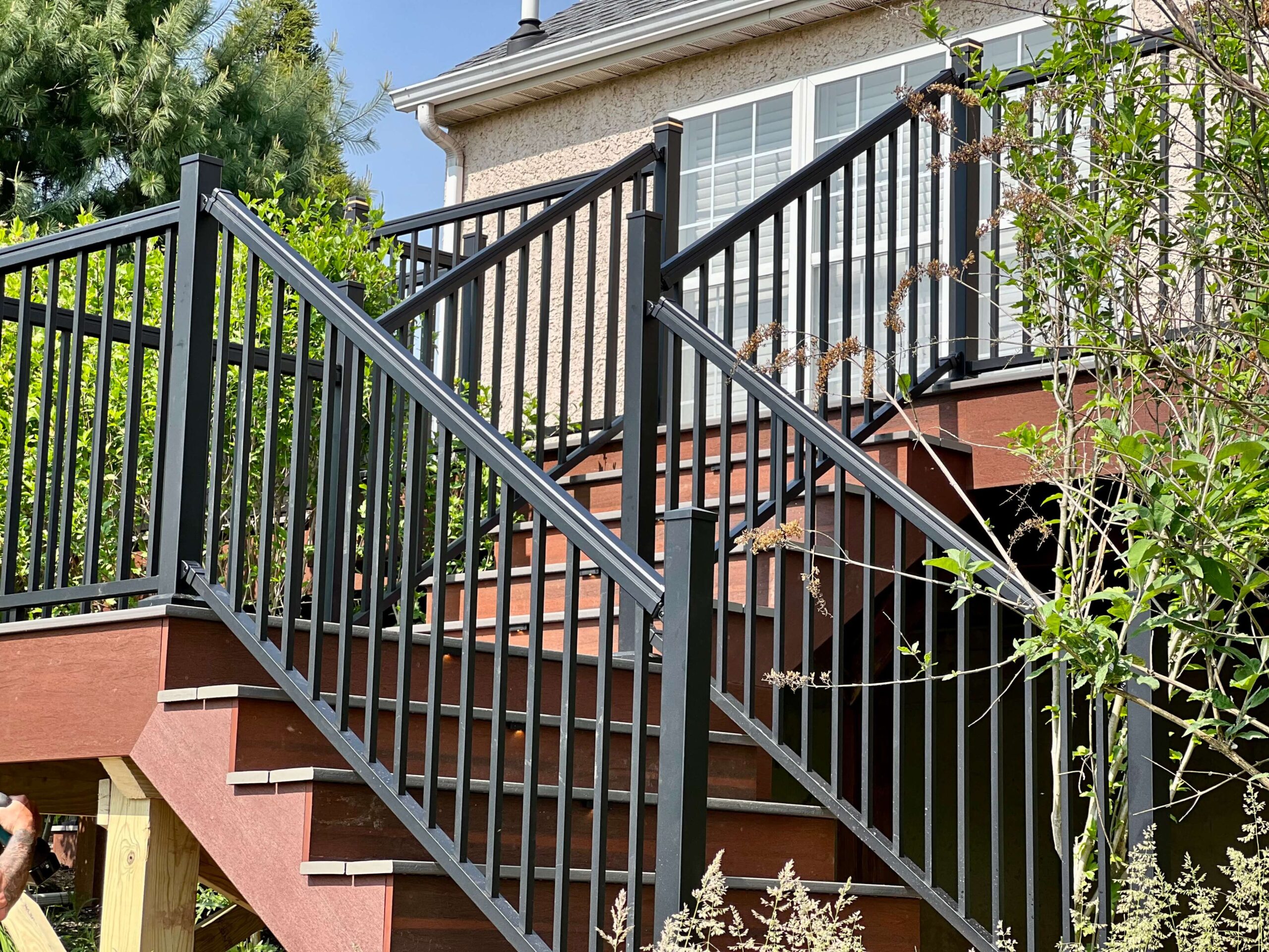 A multi-level wooden deck with wide stairs, featuring sleek black metal railings, surrounded by greenery and attached to a stucco house with large windows - Metal Railing ideas - Deck Railings