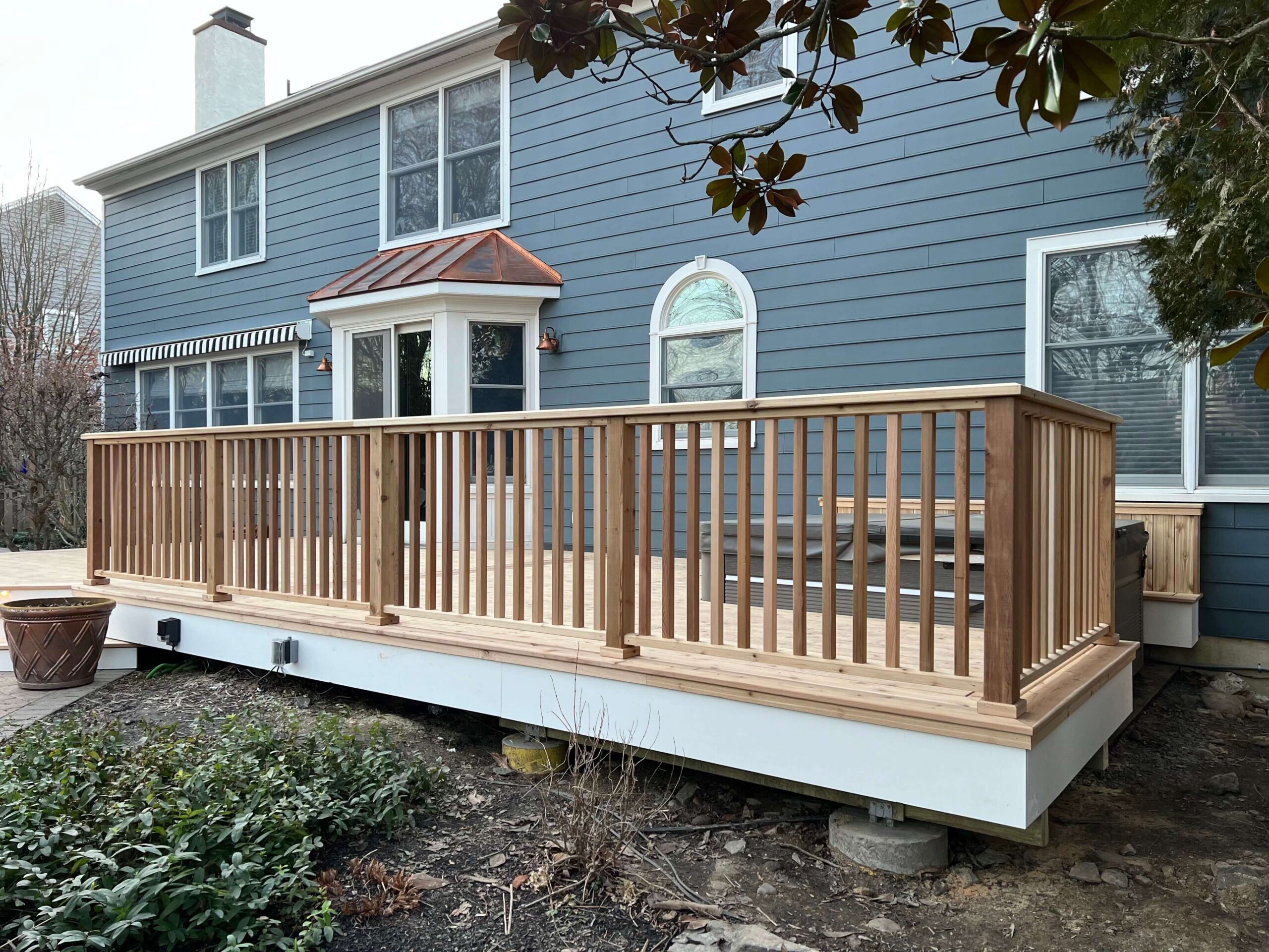 An elevated natural wood deck with vertical wooden railings and white trim, attached to a blue house with large windows and copper roofing accents - wood deck railings contractors - Deck Railings ideas