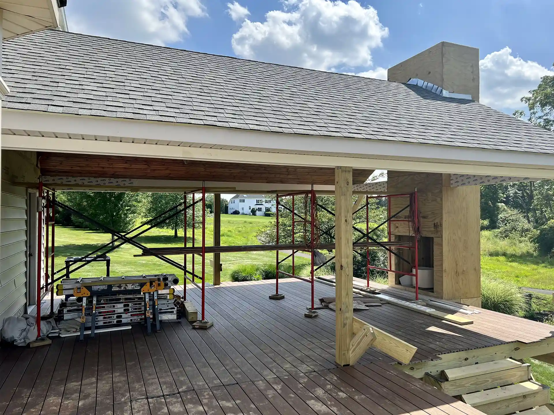 Partially constructed covered deck with brown composite flooring, wooden posts, and scaffolding, set in a scenic backyard