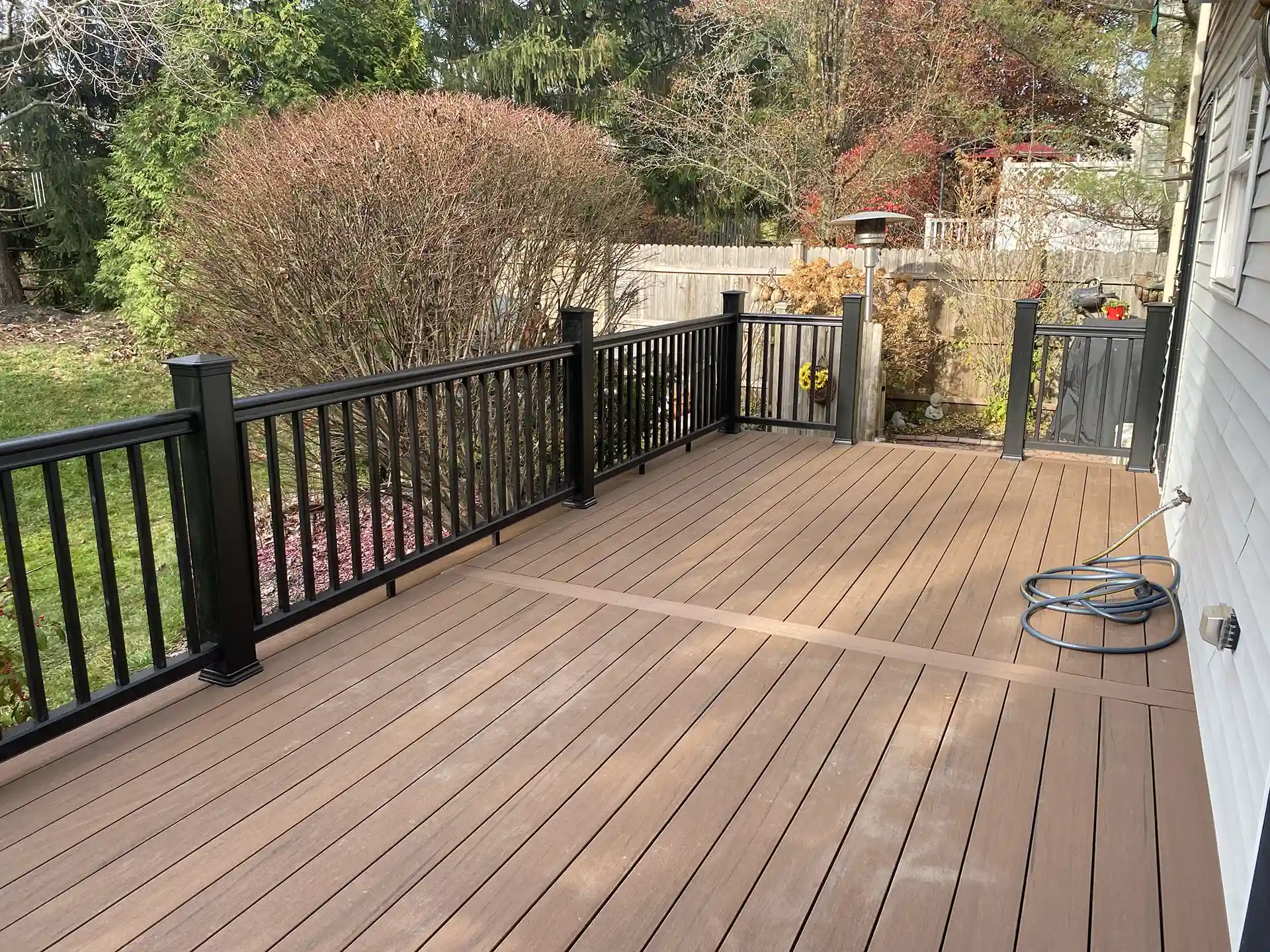 Brown composite deck with black railing, surrounded by a landscaped backyard with trees and shrubs.