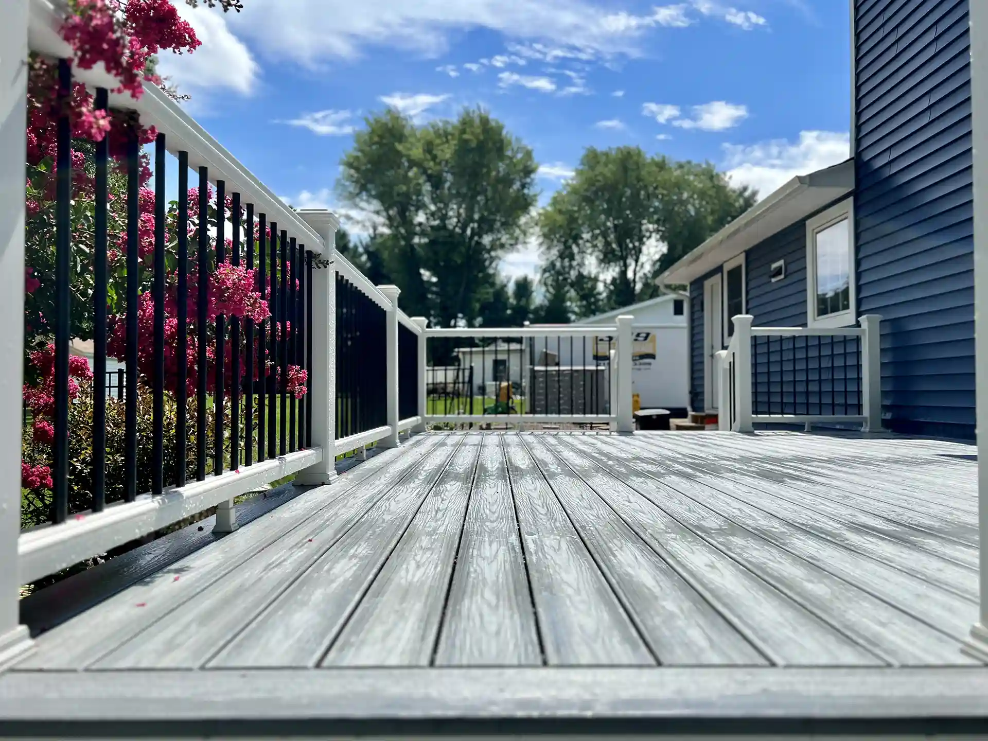 Spacious gray composite deck with white and black railing, attached to a blue house, surrounded by greenery and blooming flowers.