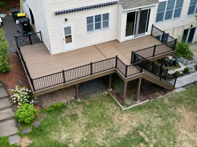 Large multi-level composite deck with black metal railings, attached to a beige siding house, elevated with strong wood support posts, and a small landscaped backyard area.