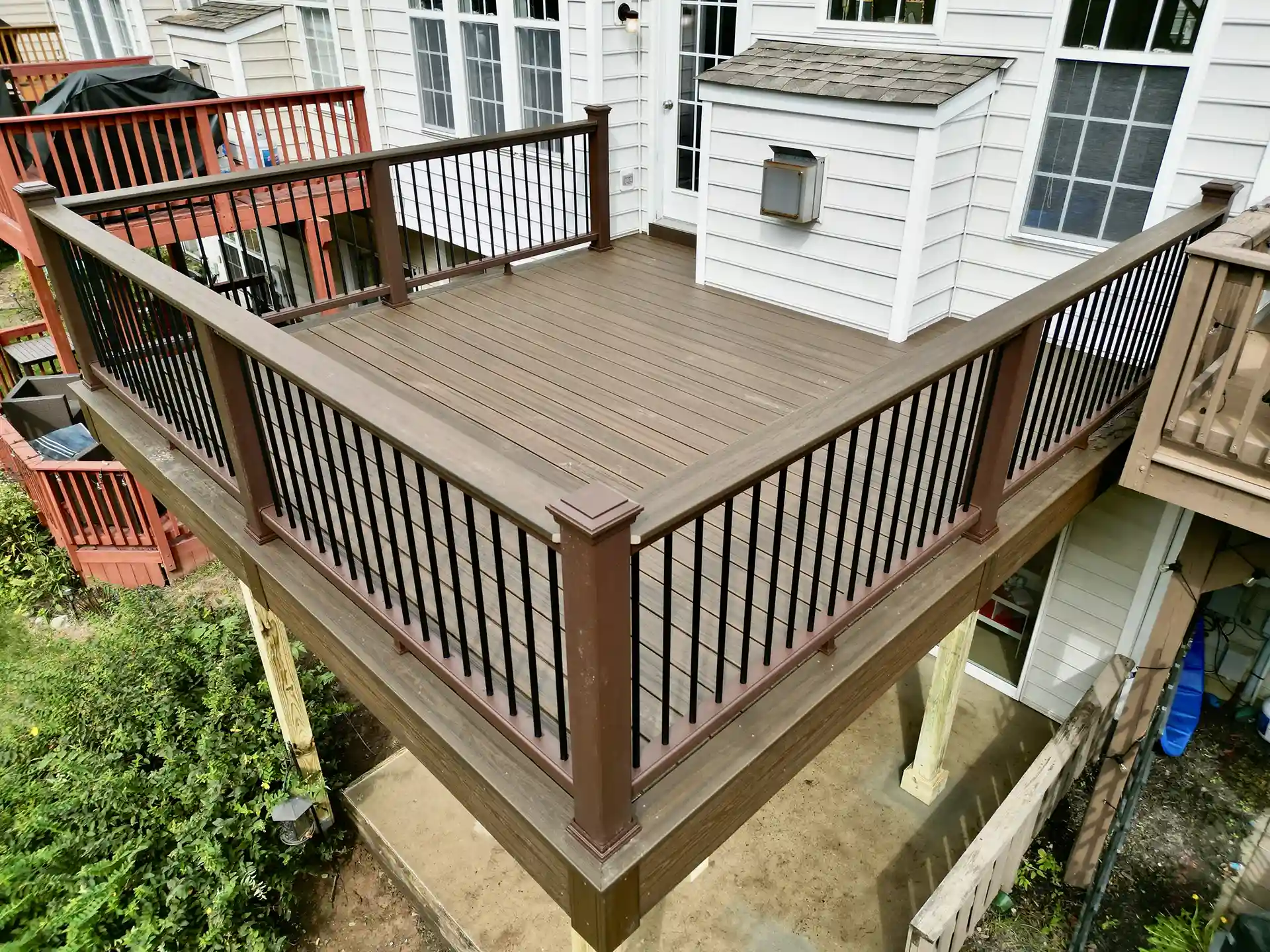 Elevated brown composite deck with black metal baluster railings, attached to a white siding house, supported by pressure-treated wood posts. 