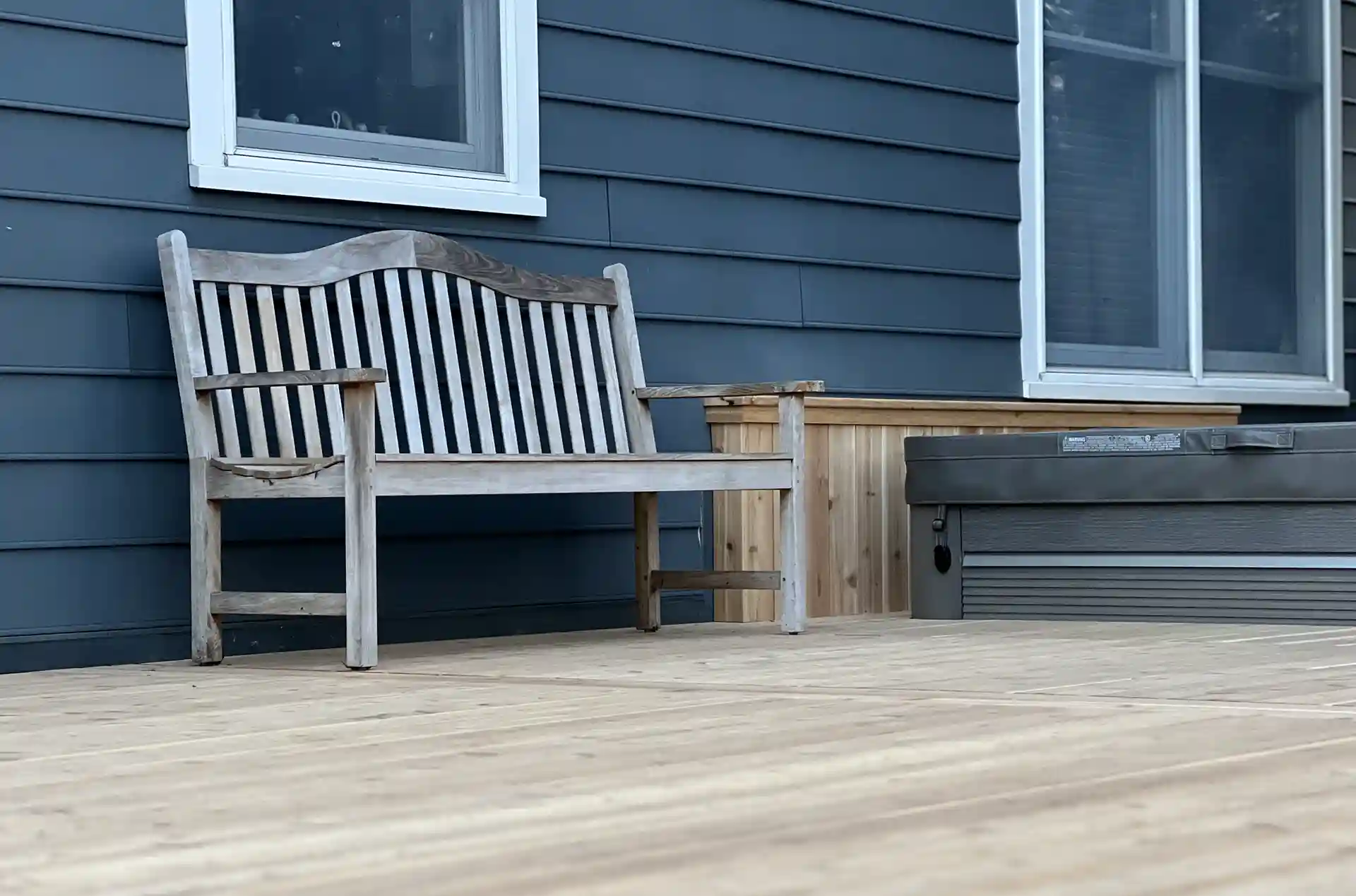 A wooden bench and hot tub positioned on a newly installed deck, with a gray exterior wall and large windows in the background. 