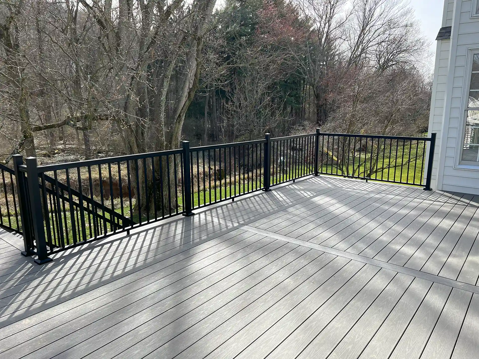 A spacious deck featuring gray composite decking with black metal railings, overlooking a green yard with trees in the background. 