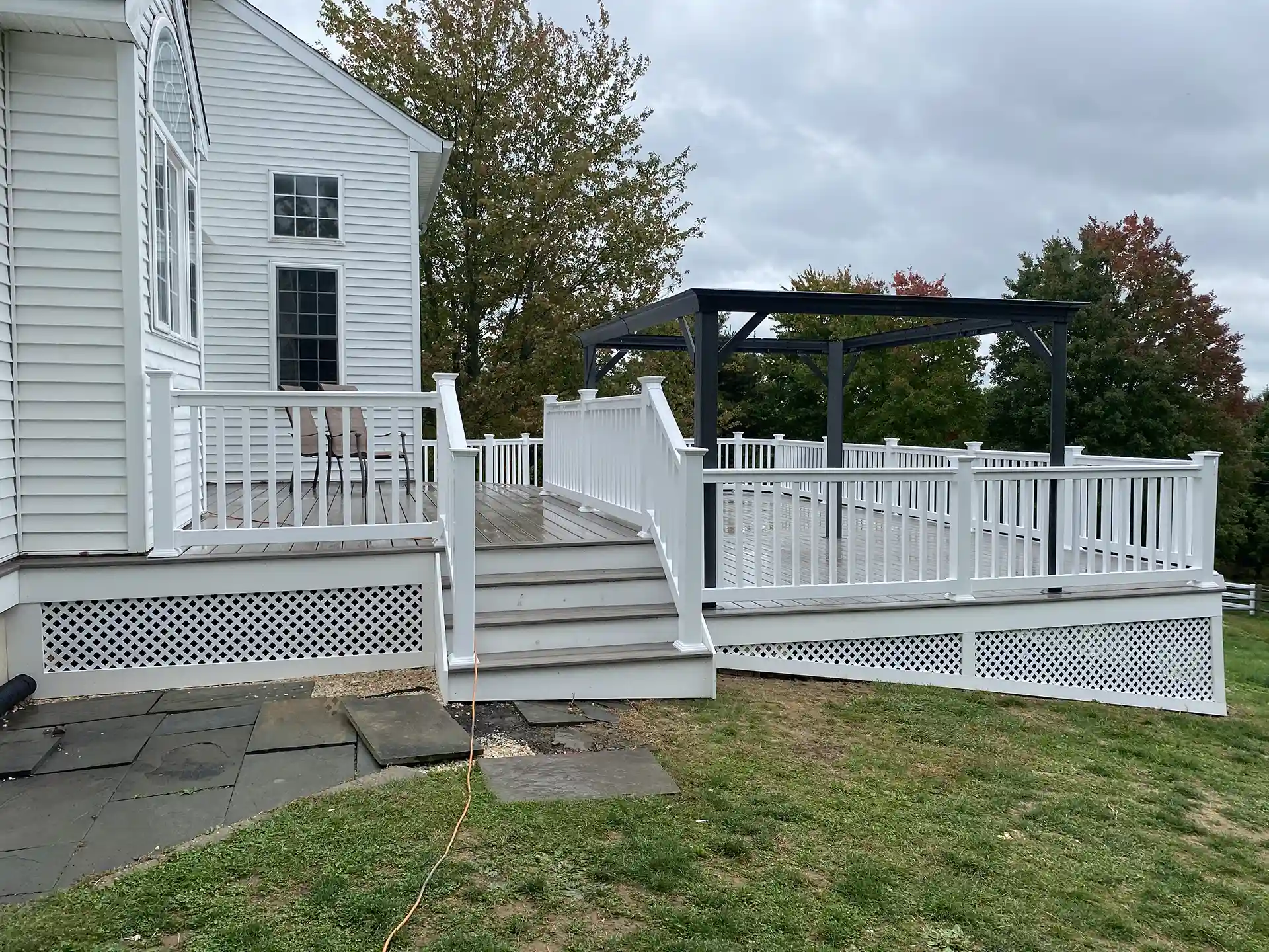 A white deck with a staircase, featuring white railing and lattice skirting, leading to a large deck area with a black pergola in the background. 