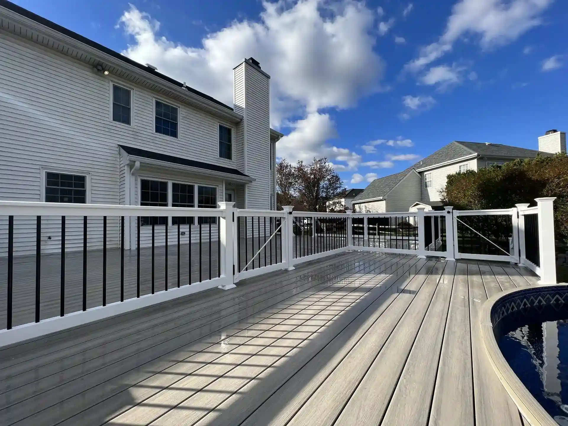 Deck with white and black railing with gray composite decking. The background features a blue sky with clouds and a pool on the right side of the deck. 