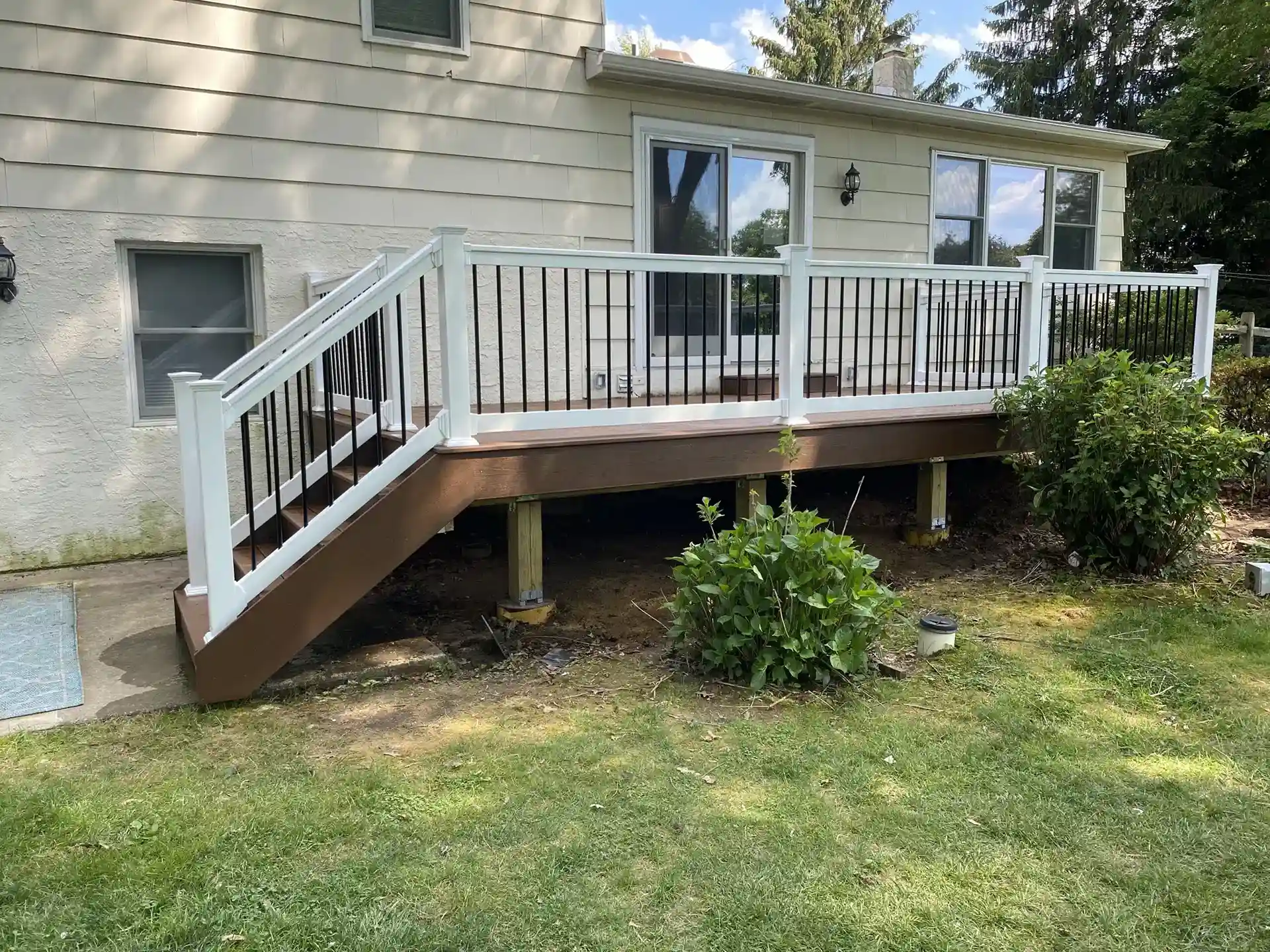 Deck with white and black railing, featuring a brown composite deck. The deck is elevated with visible support beams beneath, and the background includes a beige house with large windows and lush greenery. 